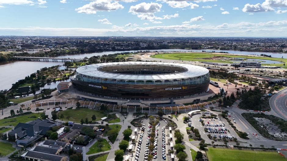 A wide aerial view of the exterior of the Optus Stadium, located near Twickenham Stadium, with lush landscaped gardens and pathways in the foreground. The stadium's modern, oval structure features a p